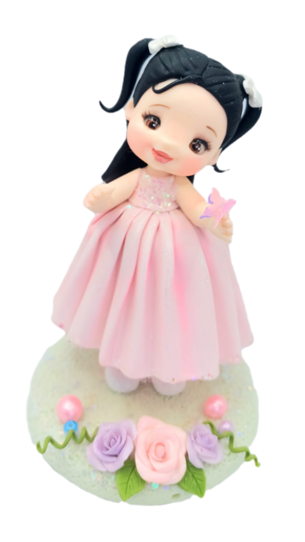 Figura decorativa de niña con vestido rosa, hecha a mano, con detalles florales y base decorada. Ideal para decoraciones de pasteles y eventos.